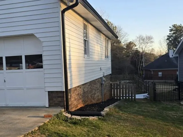 New black gutters and downspout tied into a corrugated drain line on a white West Georgia house
