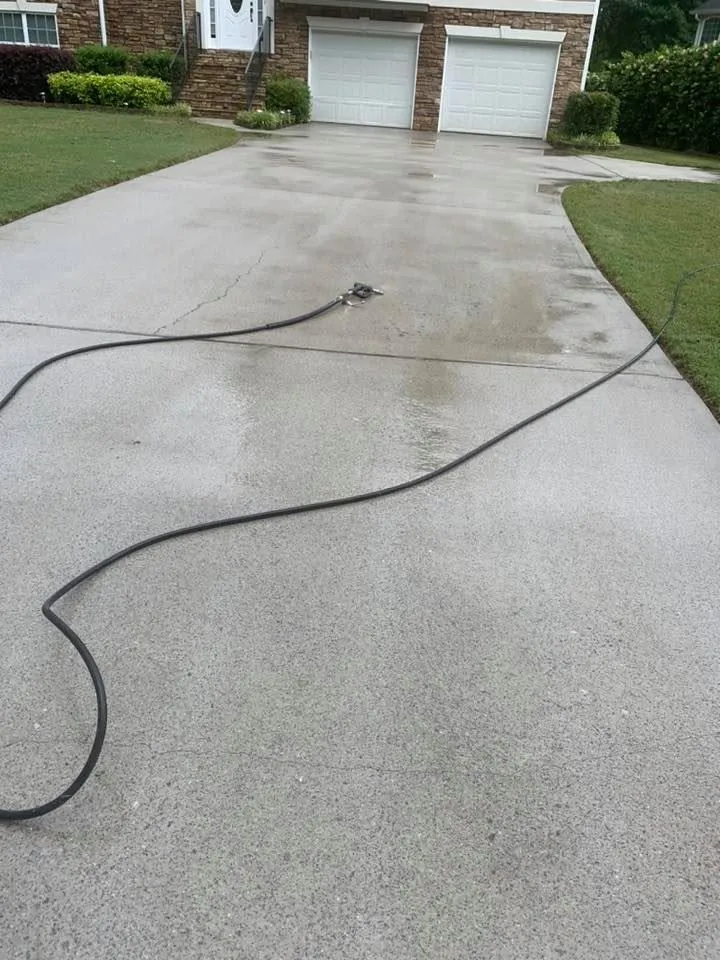 Pressure washer hose snaking across a wet light-grey concrete driveway to a wand resting mid-slab, stone-accent garage above