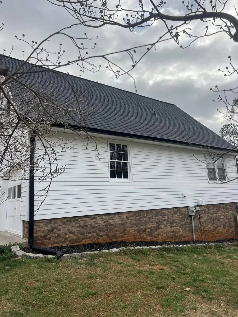 White farmhouse-style home with black gutters along a dark shingle roofline and a black downspout at the brick-skirted corner