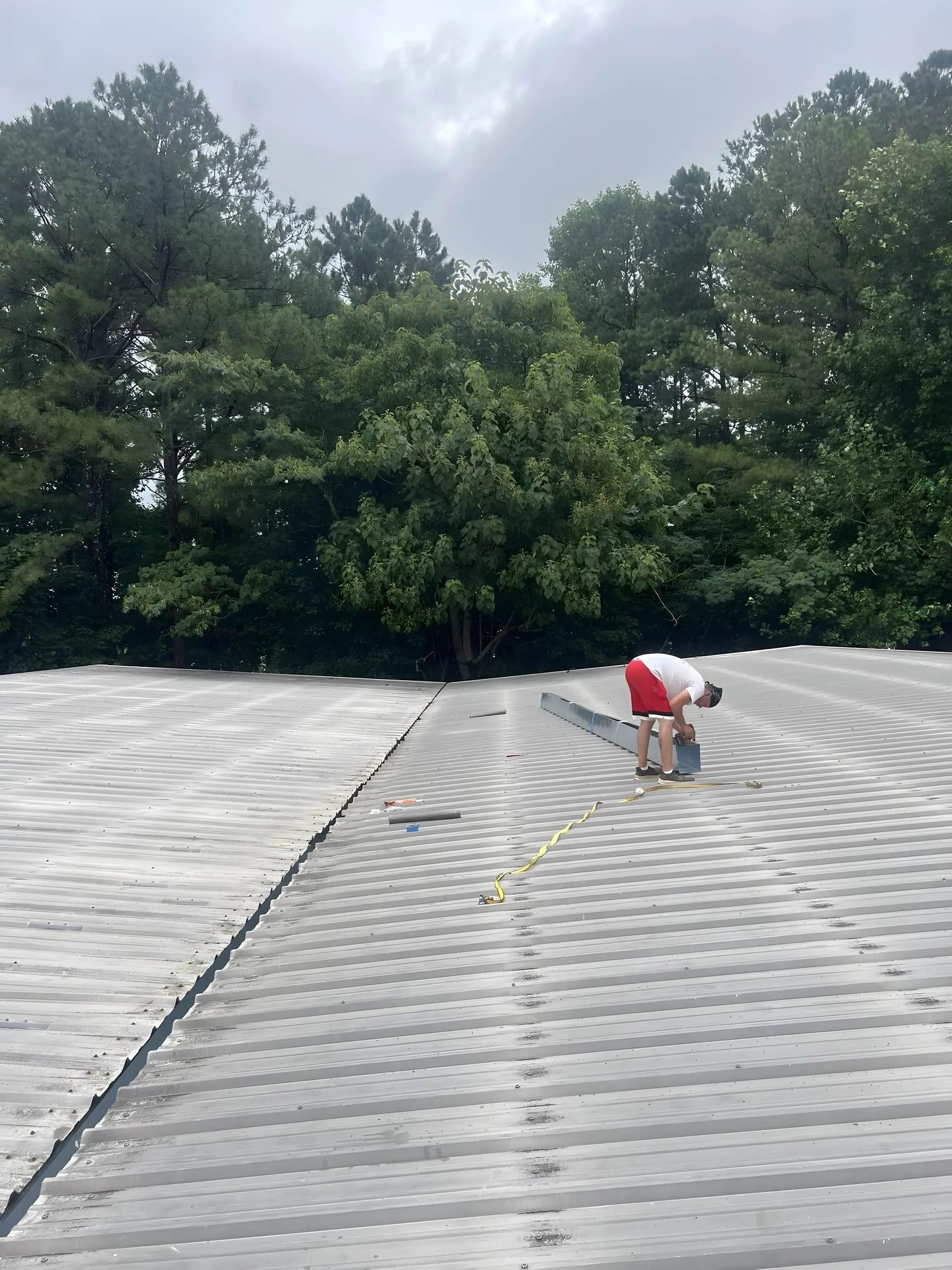 Crew member kneeling at the gutter edge of a tall metal roof, hanging a seamless gutter run, green trees behind
