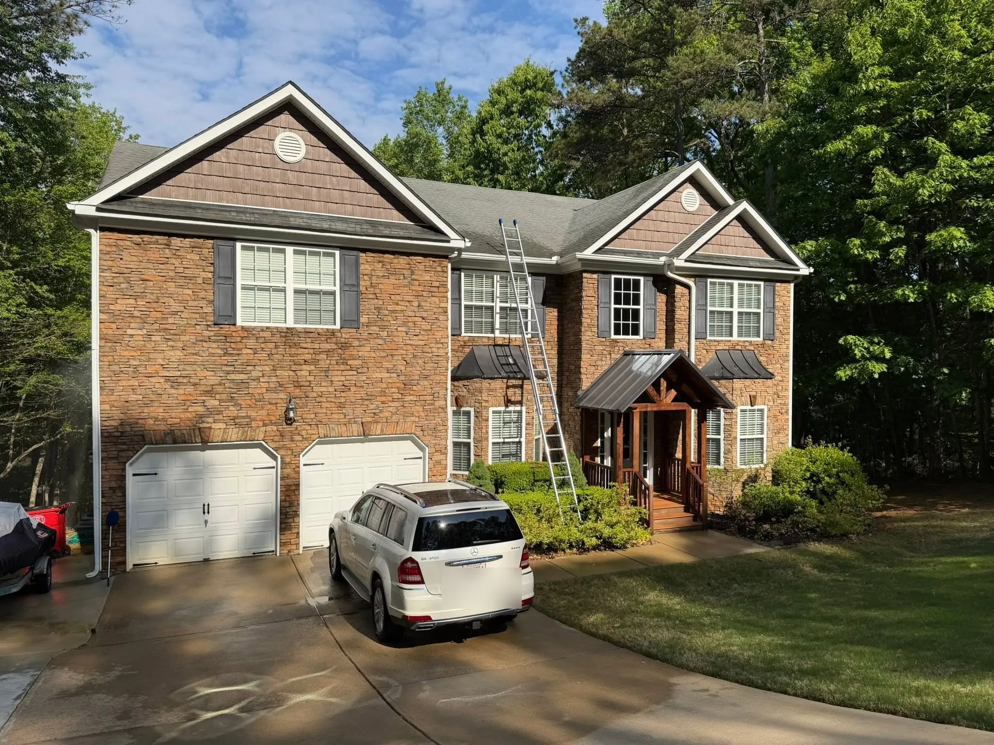 A large stone-and-shake two-story home on a wooded lot with fresh white gutters, a ladder set for the gutter work.