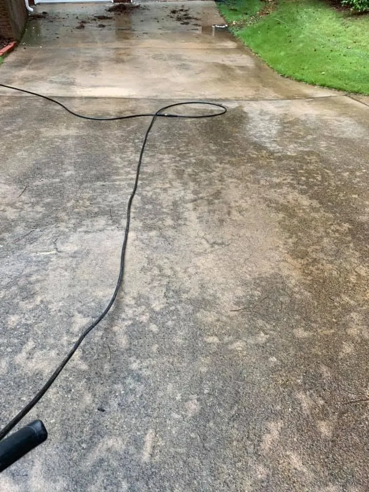 Wet concrete driveway mid-rinse with a pressure washer hose and wand lying across the slab, garage doors above