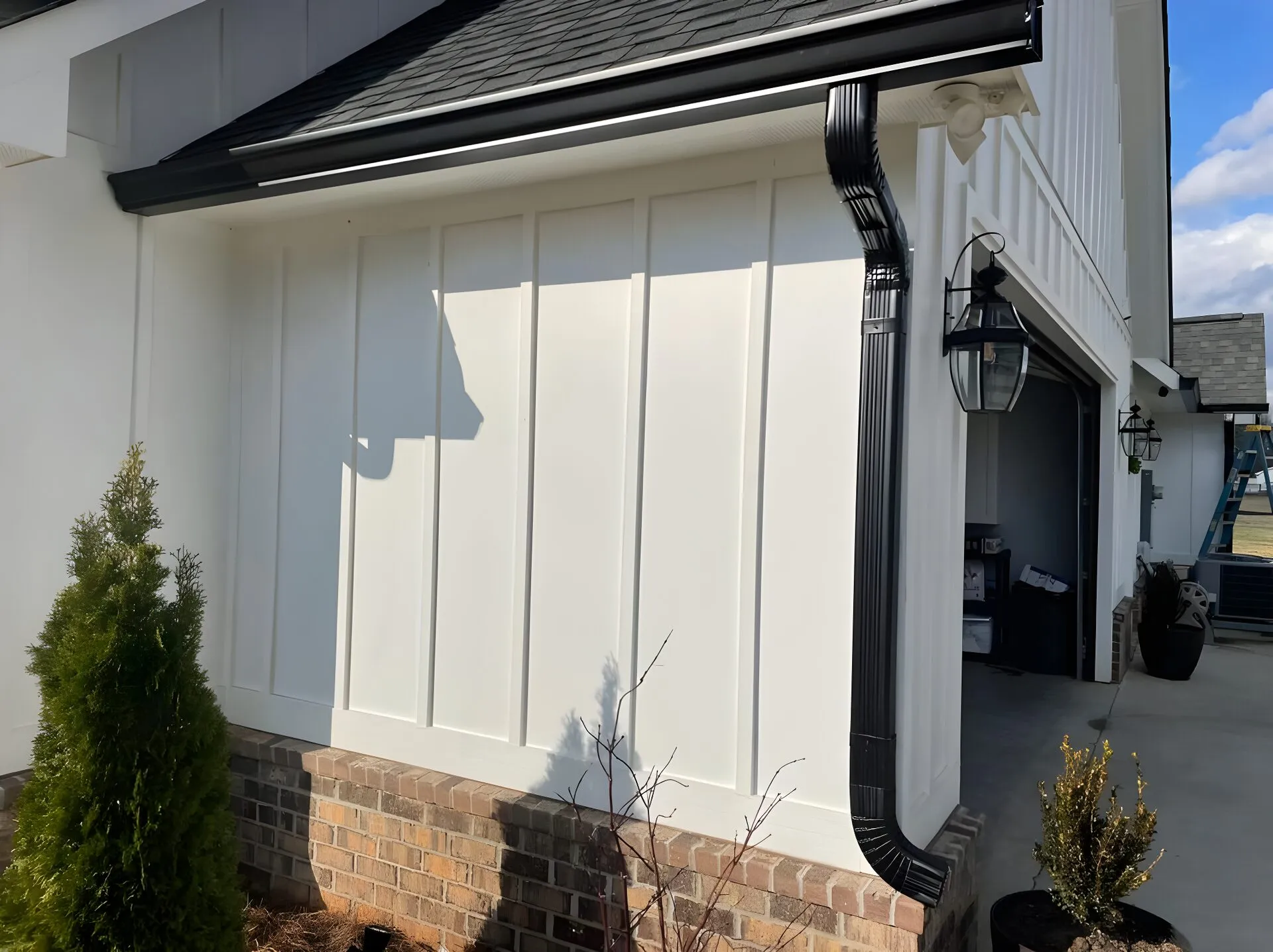 Black seamless gutter and downspout on a white board-and-batten farmhouse corner, brick base and blue sky